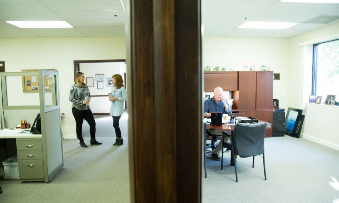 two female employees discussing work at their desks separated by a wall where a male is writing on papers in his office