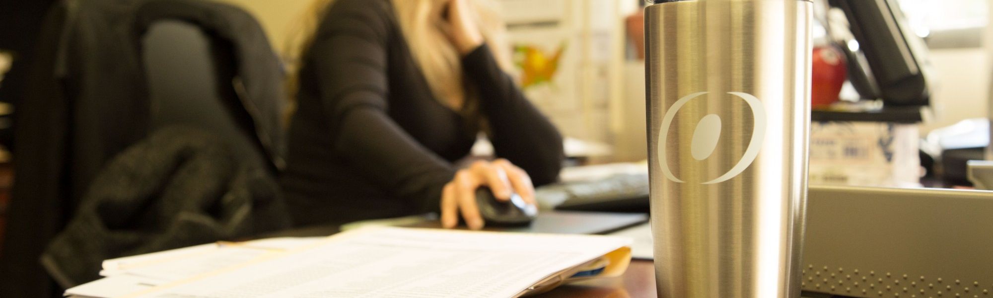 Woman typing at her desk top computer with an O'Brien branded thermos. The thermos displays the O'Brien