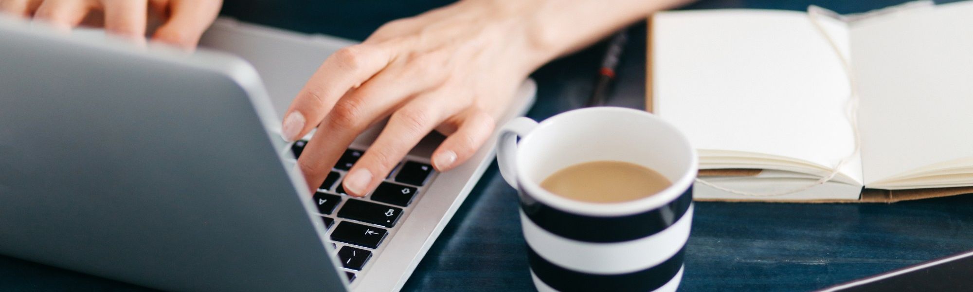 Woman typing on a laptop next to a cup full of coffee