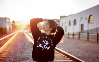 Girl with her hands on her head. Sun shining behind her on train tracks. 